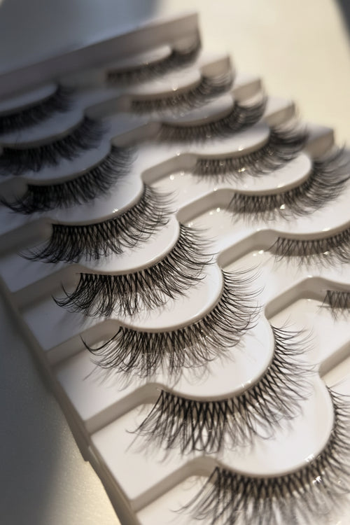 A close-up of several false eyelashes arranged in a white plastic tray, photographed on a light surface with soft shadows.